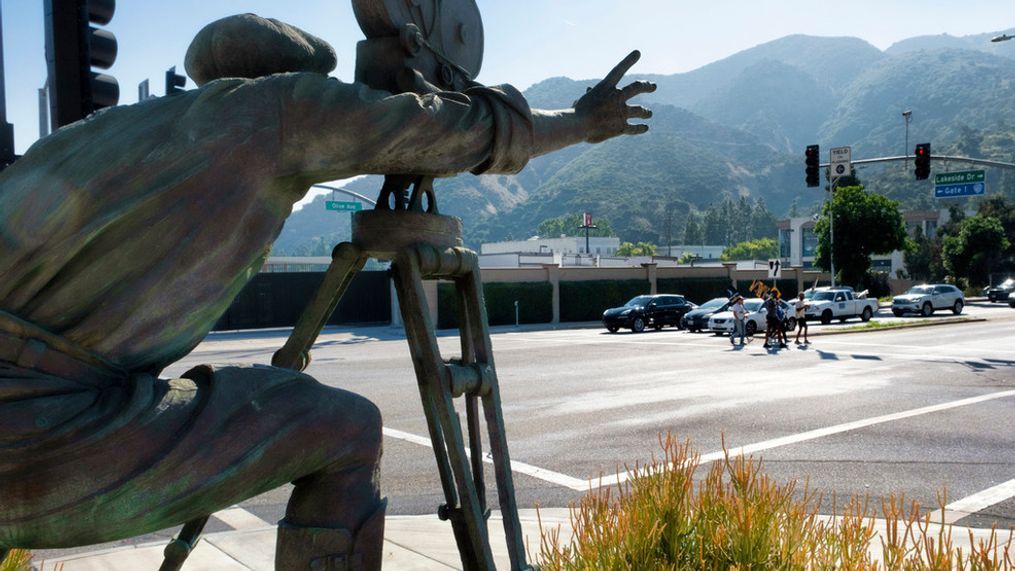 With a statue of a cameraman in the foreground, SAG-AFTRA picketers carrying signs cross a street near the gates of Warner Bros. studios in Burbank, Calif., Tuesday, Sep. 26, 2023. (AP Photo/Richard Vogel)