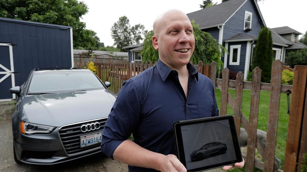 In this June 27, 2018, photo, Christian Kingery poses for photo at his home in Preston, Wash., near Seattle, next to the leased Audi he is driving until he can take delivery of a long-awaited Tesla Model 3, which is shown on his tablet computer. Kingery, who is on an official waiting list for his car, says Tesla has told him it could take until between September and November of 2018 for his car to be delivered, but he fears it might be even longer. (AP Photo/Ted S. Warren)