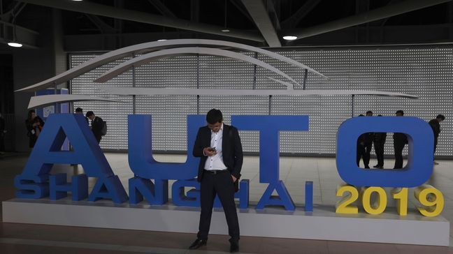 A man waits near a sign for the Auto Shanghai 2019 show in Shanghai Tuesday, April 16, 2019. Automakers are showcasing electric SUVs and sedans with more driving range and luxury features at the Shanghai auto show, trying to appeal to Chinese buyers in their biggest market as Beijing slashes subsidies that have propelled demand. (AP Photo/Ng Han Guan)